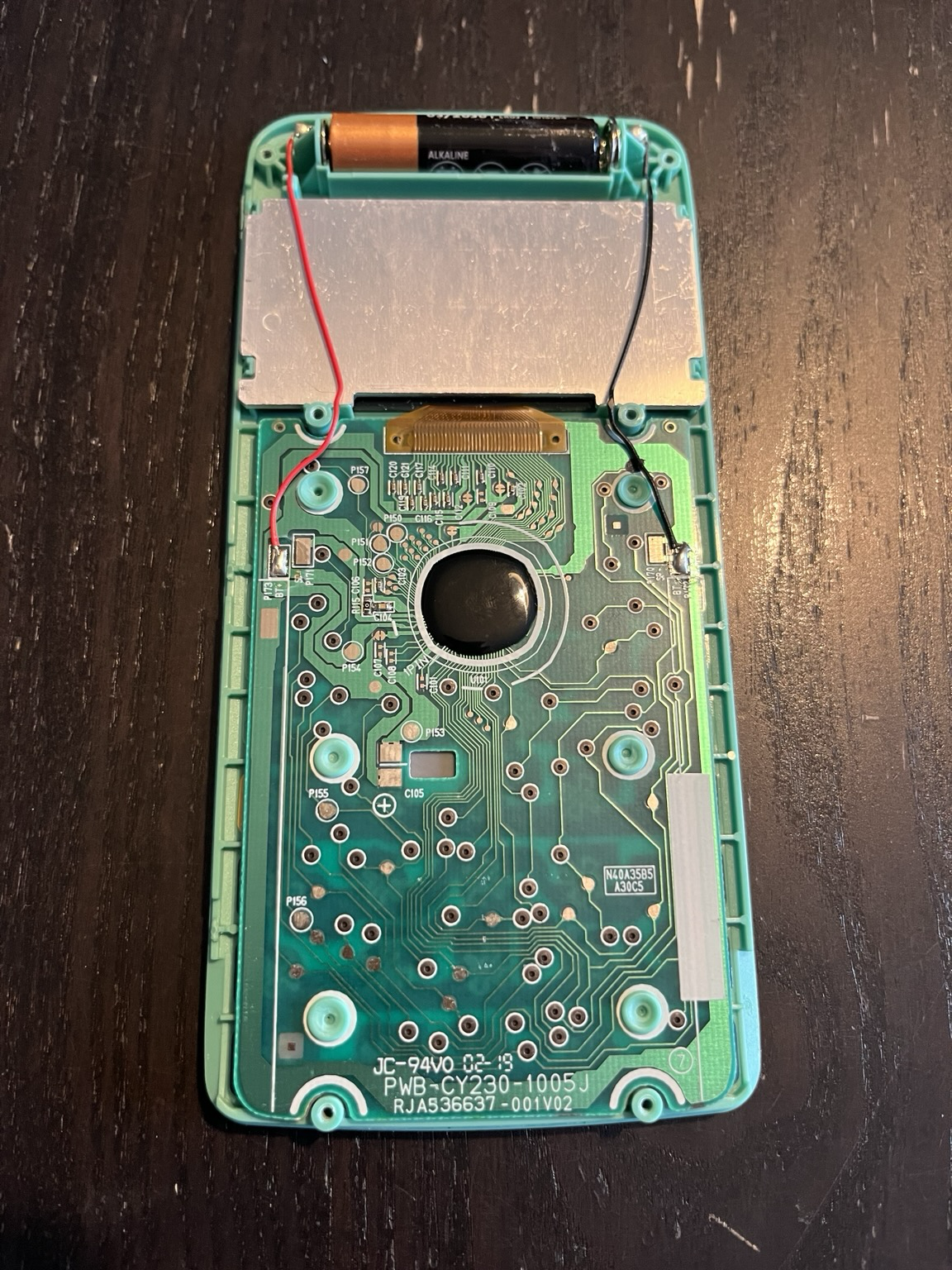 This is the picture of the backside the green motherboard of a Casio fx-92B calculator. From top to bottom, the AAA battery is on top of the picture, the backside of the LCD screen is visible, then comes the motherboard. The CPU is hidden behind a drop of black epoxy (~1.5cm diameter). Molten plastic rivets are holding the motherboard close to the plastic chassis, with the keyboard in between.